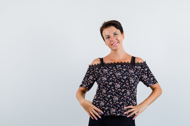 Mulher madura de mãos dadas na cintura em uma blusa floral e saia preta e parecendo alegre. vista frontal.