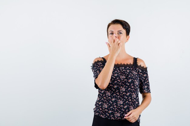 Mulher madura em blusa floral e saia preta puxando o nariz para cima com os dedos e olhando séria, vista frontal.