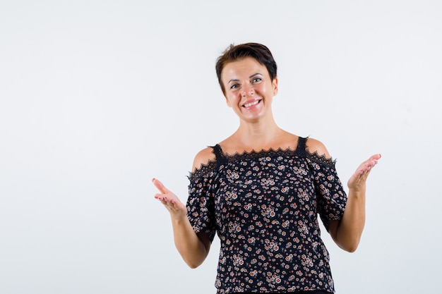 Mulher madura em blusa floral, saia preta, esticando as mãos de forma questionadora e olhando alegre, vista frontal.