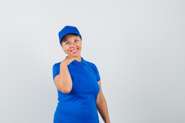 Mulher madura em t-shirt azul, apoiando o queixo na mão e olhando feliz.