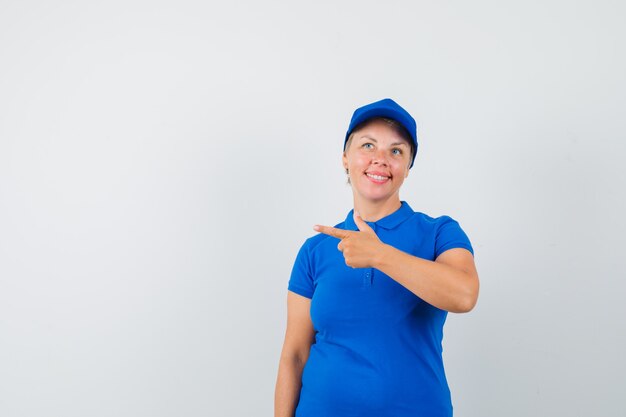 Mulher madura em t-shirt azul apontando para o lado esquerdo e parecendo alegre.