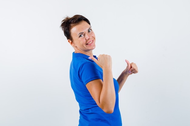 Mulher madura em t-shirt azul apontando para trás com os polegares e parecendo confiante.