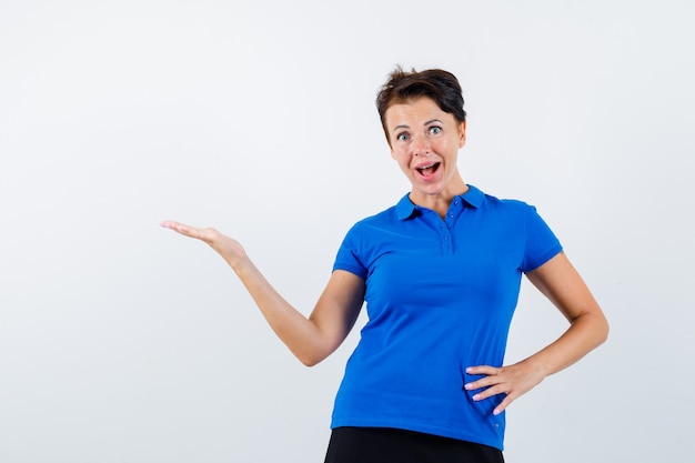 Mulher madura em t-shirt azul fazendo gesto de boas-vindas e olhando espantado, vista frontal.