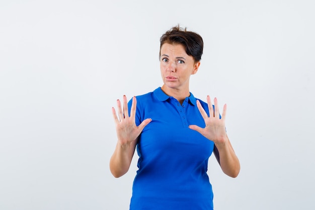 Mulher madura em t-shirt azul, mostrando o gesto de parar e olhando com medo, vista frontal.