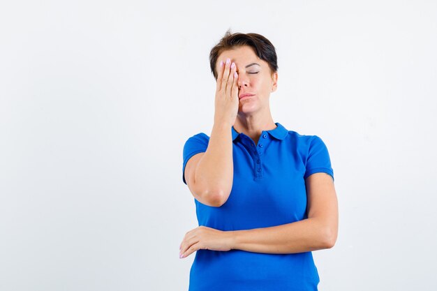 Mulher madura em t-shirt azul, segurando a mão no olho e olhando pensativa, vista frontal.