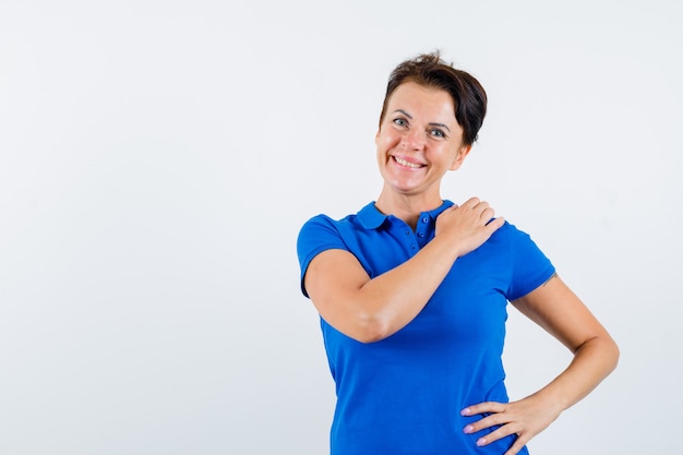 Mulher madura em t-shirt azul, segurando a mão no ombro e olhando feliz, vista frontal.