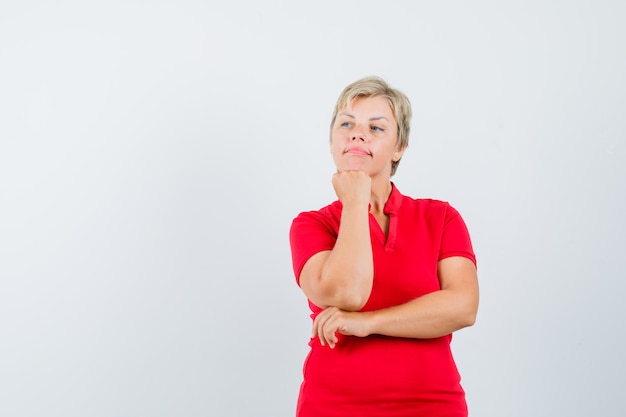 Mulher madura em t-shirt vermelha, apoiando o queixo no punho levantado e olhando pensativa.