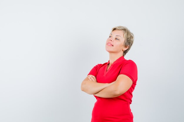 Mulher madura em uma camiseta vermelha em pé com os braços cruzados e parecendo pensativa