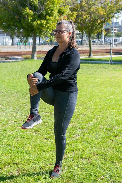 Mulher madura focada em roupas de fitness, fazendo exercícios matinais na grama do parque, em pé e esticando as pernas. Conceito de bem-estar ou estilo de vida ativo