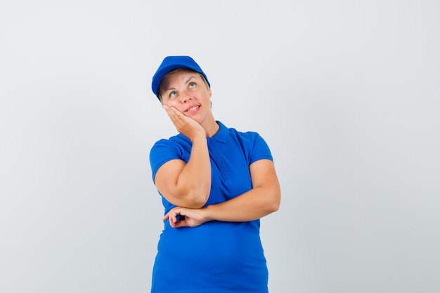 Mulher madura, inclinando a bochecha na palma da mão levantada em uma camiseta azul e parecendo esperançosa.