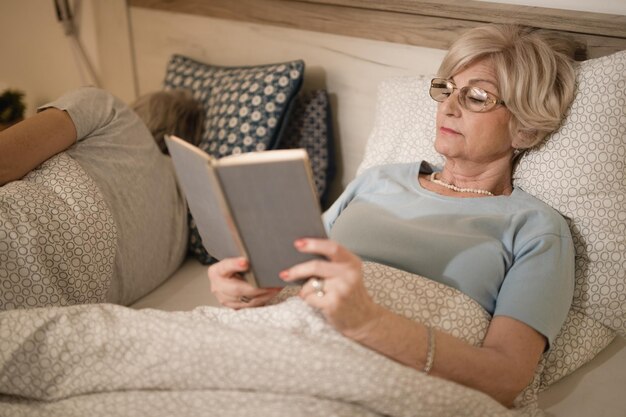 Mulher madura lendo livro enquanto deitada na cama à noite Seu marido está dormindo ao lado dela