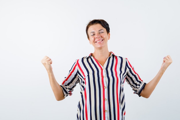 Mulher madura mostrando o gesto do vencedor, fechando os olhos em uma camisa listrada e parecendo com sorte. vista frontal.