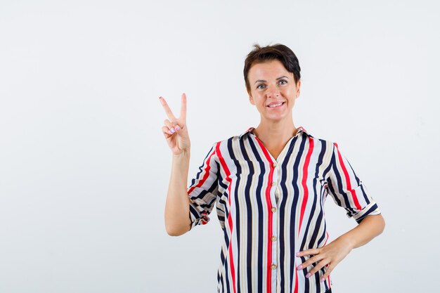 Mulher madura mostrando um gesto de paz, segurando a mão na cintura em uma camisa listrada e olhando confiante, vista frontal.