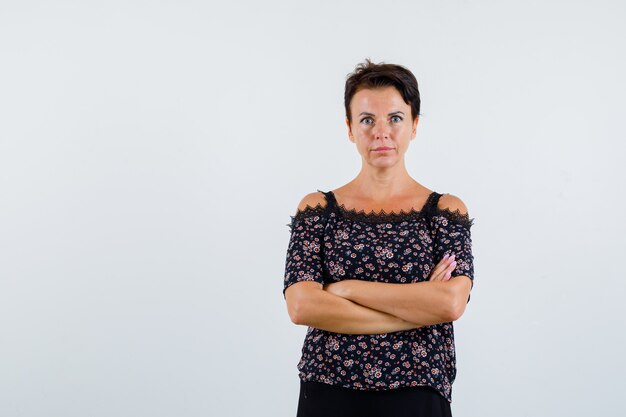 Mulher madura na blusa floral e saia preta em pé com os braços cruzados e olhando séria, vista frontal.