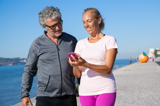 Mulher madura positiva usando o aplicativo de fitness no celular depois de correr, mostrando a tela para o homem. comunicação e gadget para o conceito de esporte