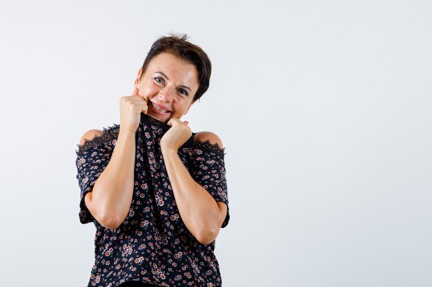 Mulher madura puxando o colarinho no queixo em blusa floral, saia preta e olhando alegre, vista frontal.