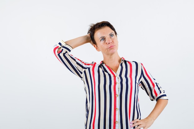 Mulher madura, segurando a mão na cintura, segurando uma mão na cabeça em uma blusa listrada e olhando pensativa, vista frontal.