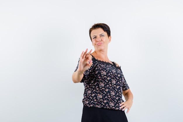 Mulher madura segurando uma mão na cintura, mostrando sinal de ok na blusa floral e saia preta e parecendo confiante. vista frontal.