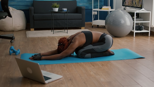 Mulher magra de pele negra sentada no mapa de ioga, fazendo exercícios de ginástica matinal na sala de estar, enquanto assiste a um vídeo aeróbico online usando um laptop