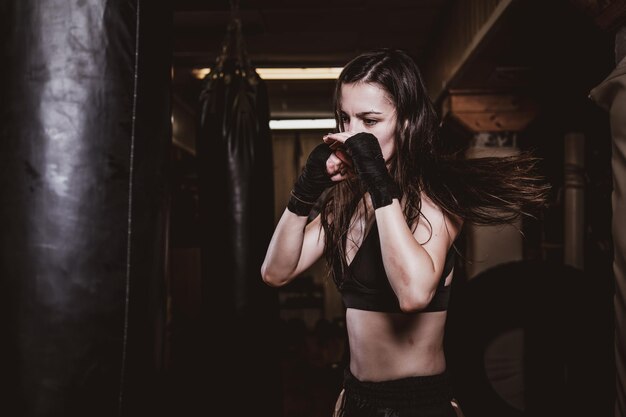 Mulher magra focada tem um treinamento de boxe com saco de pancadas no ginásio escuro.