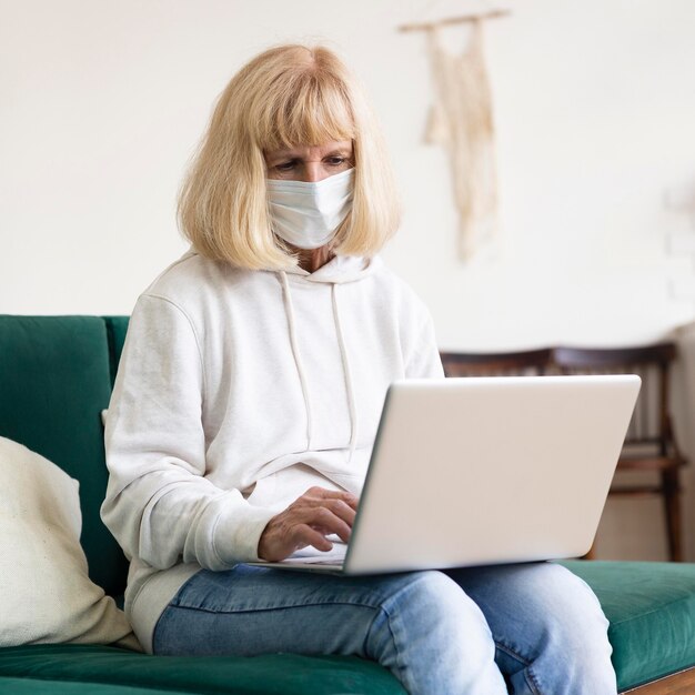 Mulher mais velha trabalhando em um laptop em casa, usando máscara médica