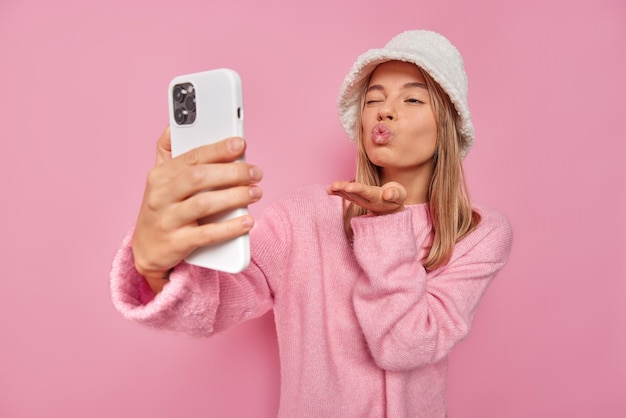 mulher manda beijo no ar na câmera do celular tira foto glamourosa de selfie pisca os olhos e faz beicinho usa chapéu e jumper isolado no rosa gosta de chat por vídeo com o namorado.