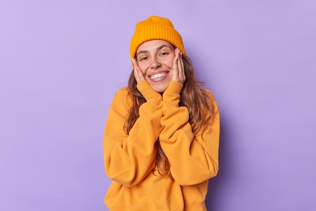 mulher mantém as mãos nas bochechas sorrisos dentuços tem expressão alegre vestida de jumper casual laranja e chapéu isolado no roxo. Linda garota milenar se sentindo muito feliz