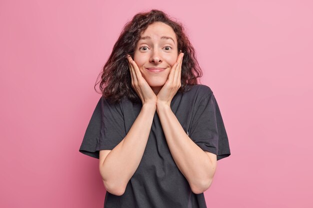 mulher mantém as mãos nas bochechas sorrisos reage agradavelmente a notícias incríveis vestida com uma camiseta preta casual isolada rosa