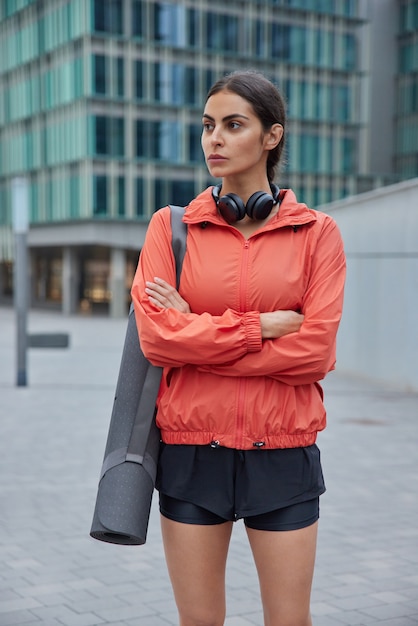 Foto grátis mulher mantém os braços cruzados olhando para longe fazendo exercícios no tapete de borracha de fitness ao ar livre vestida com roupas esportivas, pronta para o treino