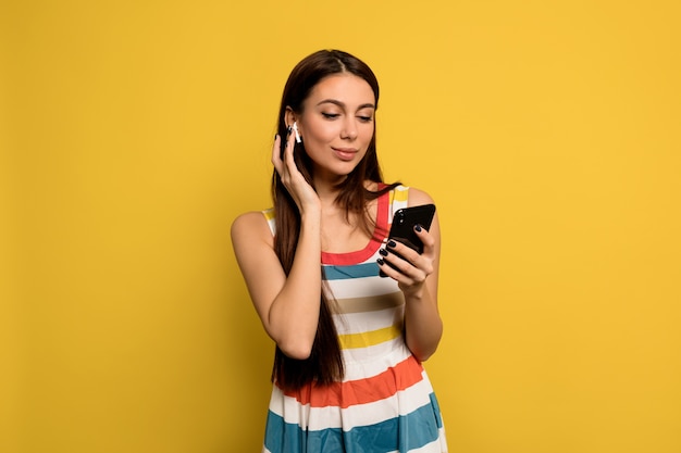 Mulher maravilhosa com cabelo comprido e maquiagem nua usando um vestido brilhante ouvindo música e segurando o smartphone