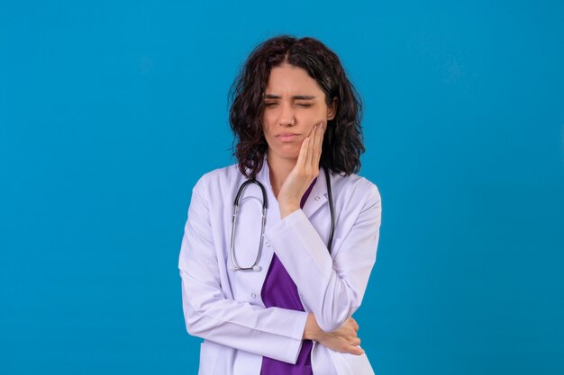 mulher médica vestindo jaleco branco com estetoscópio em pé com a mão na bochecha, forma de dor de dente