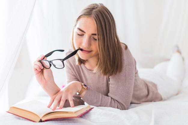 Mulher, mentindo cama, morder, pretas, óculos, em, boca, enquanto, leitura, livro