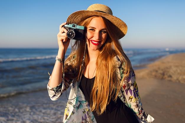 Mulher mesquinha feliz com longos cabelos loiros se divertindo e tirando fotos na praia perto do oceano com uma câmera vintage, cores ensolaradas