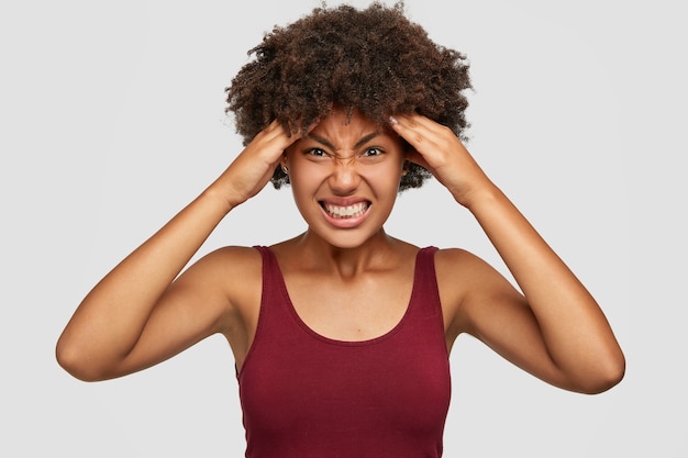 Mulher miserável de pele escura sente desconforto, sofre de dor de cabeça, não consegue focar, cerra os dentes e franze a testa