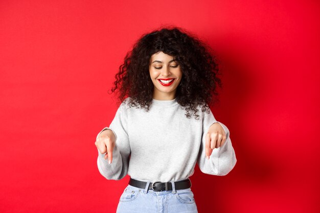 Mulher moderna com penteado encaracolado e lábios vermelhos apontando os dedos para baixo e sorrindo feliz mostrando adver ...