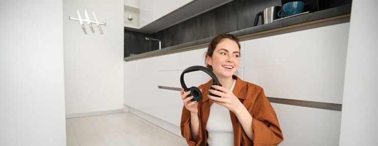 Foto grátis mulher moderna despreocupada senta-se no chão em casa sorri e olha de lado coloca fones de ouvido sem fio para
