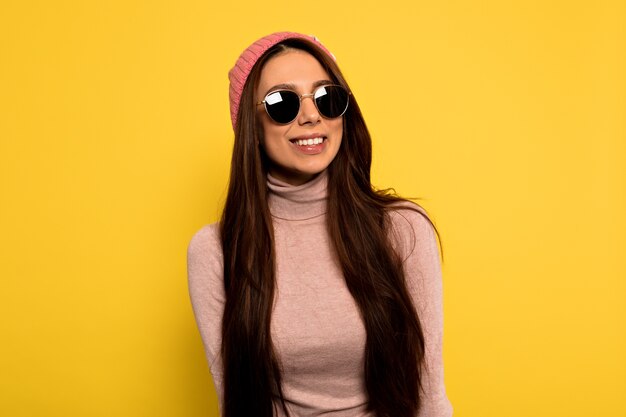 Mulher moderna e elegante hipster com longos cabelos escuros, boné rosa e óculos de sol redondos, posando com um sorriso feliz