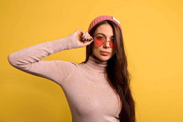 Mulher moderna elegante com cabelo longo e reto, vestindo uma camisa rosa e boné tocando seus óculos redondos rosa e sorrindo