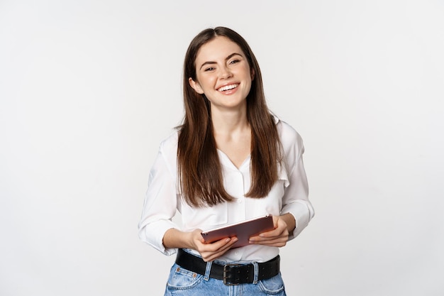 Foto grátis mulher moderna sorridente em pé com tablet digital, rindo e parecendo feliz, trabalhando, posando contra o fundo branco do estúdio.