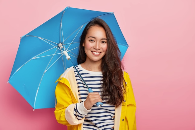 Mulher morena alegre Asain com longos cabelos escuros, usa um macacão listrado, capa de chuva amarela, segura guarda-chuva azul, faz um passeio em dia chuvoso