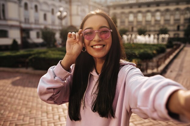 Mulher morena alegre com capuz rosa e óculos de sol da moda sorri sinceramente e tira uma selfie de bom humor lá fora