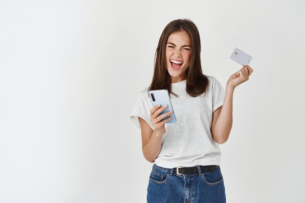 Mulher morena alegre, compras online, usando o smartphone e mostrando o cartão de crédito de plástico, sorrindo alegre na frente, parede branca.