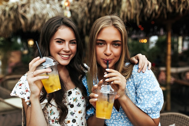 Mulher morena alegre e encaracolada em uma blusa da moda floral e uma garota loira bronzeada com top azul sorriem e segurando copos de limonada do lado de fora