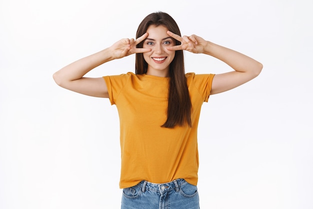 Mulher morena alegre, otimista e sortuda em uma camiseta amarela, mostrando sinal de paz ou vitória sobre os olhos, sorrindo, expressando positividade e alegria, aproveite a festa, como participar de um evento incrível, fundo branco