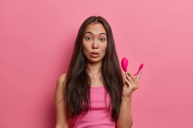 Mulher morena atônita segura vibrador inteligente moderno controlado por aplicativo remoto, visita sex shop e compra os acessórios necessários para se satisfazer, vestida casualmente, posa contra a parede rosa