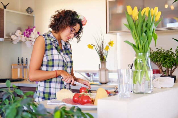 Mulher morena atraente em óculos fazendo salada de legumes na cozinha de casa.
