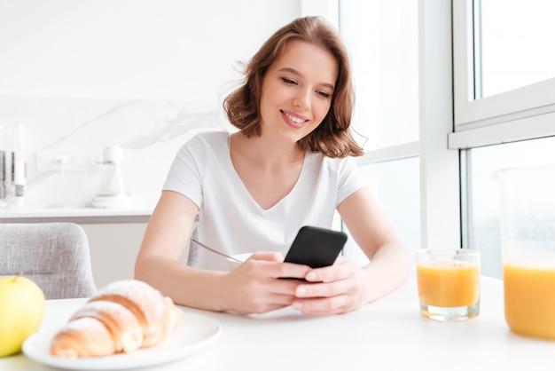 Mulher morena bonita camiseta branca usando smartphone enquanto está sentado na mesa da cozinha pela manhã