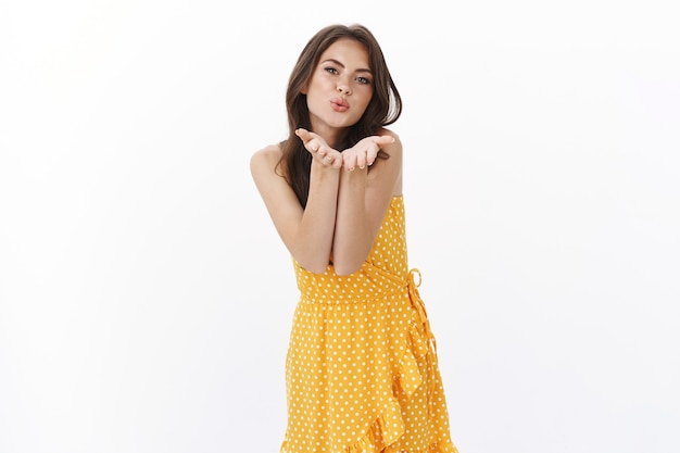 Mulher morena bonita e afetuosa em um vestido amarelo de verão, soprando beijo do vento, segure as palmas das mãos perto dos lábios de beicinho para enviar muah câmera, pareça sedutor e bobo na parede branca, clima sedutor romântico