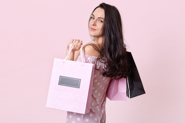 Foto grátis mulher morena bonita fica de lado, detém sacolas de compras, retorna do shopping de bom humor, posa em rosa. mulheres e o conceito de compra.