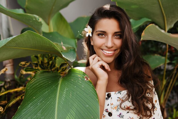 Mulher morena de cabelos compridos de ótimo humor posando com um sorriso entre as plantas tropicais